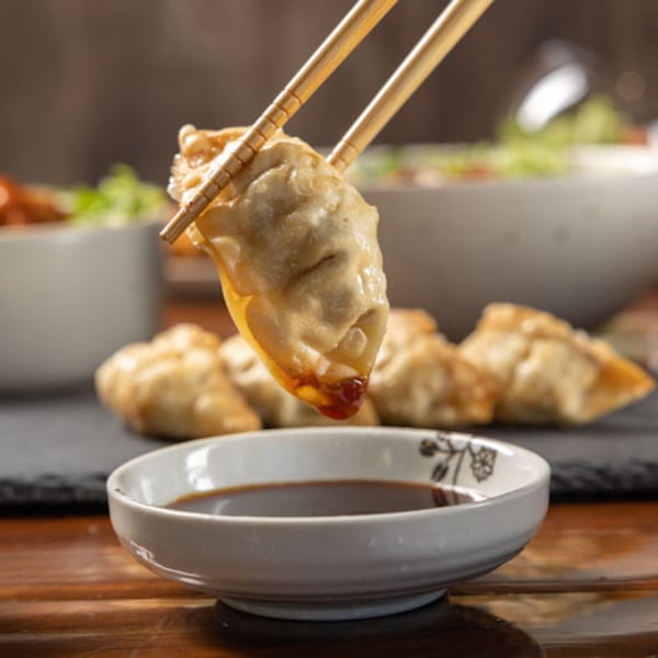 Chopsticks holding a fried Korean dumpling, dripping sauce into a small bowl.
