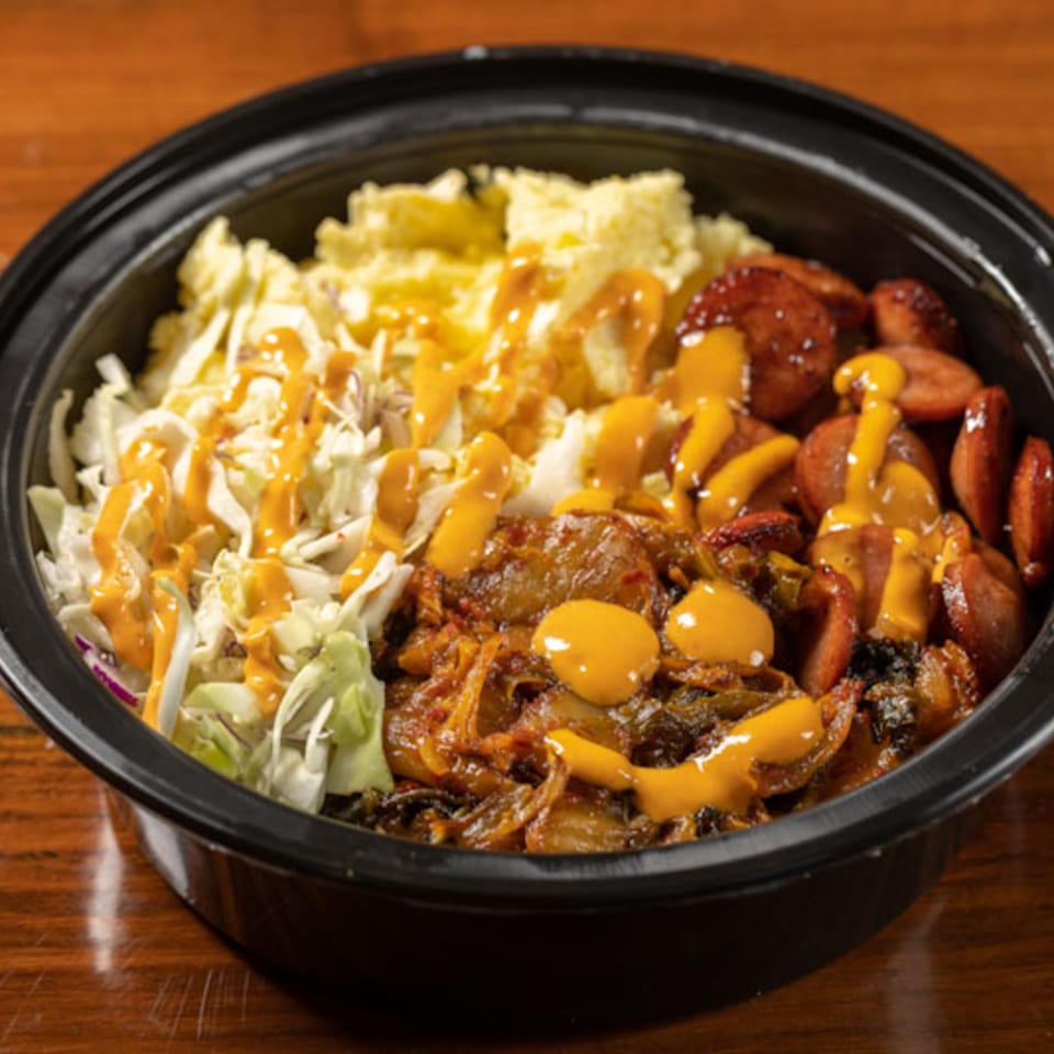 A black bowl of Korean BBQ with sausage, kimchi, coleslaw, and scrambled eggs, drizzled with sauce.