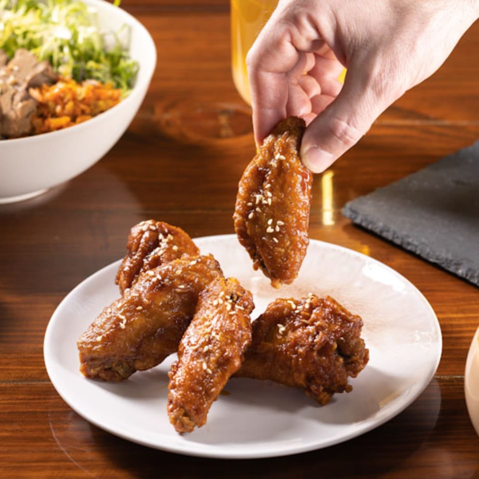 A hand picks up a sesame-sprinkled Korean fried chicken wing from a white plate on a wooden table.