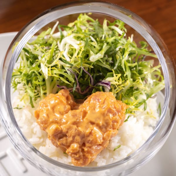 A clear bowl filled with white rice, topped with green salad and a heart-shaped portion of Korean spicy chicken.