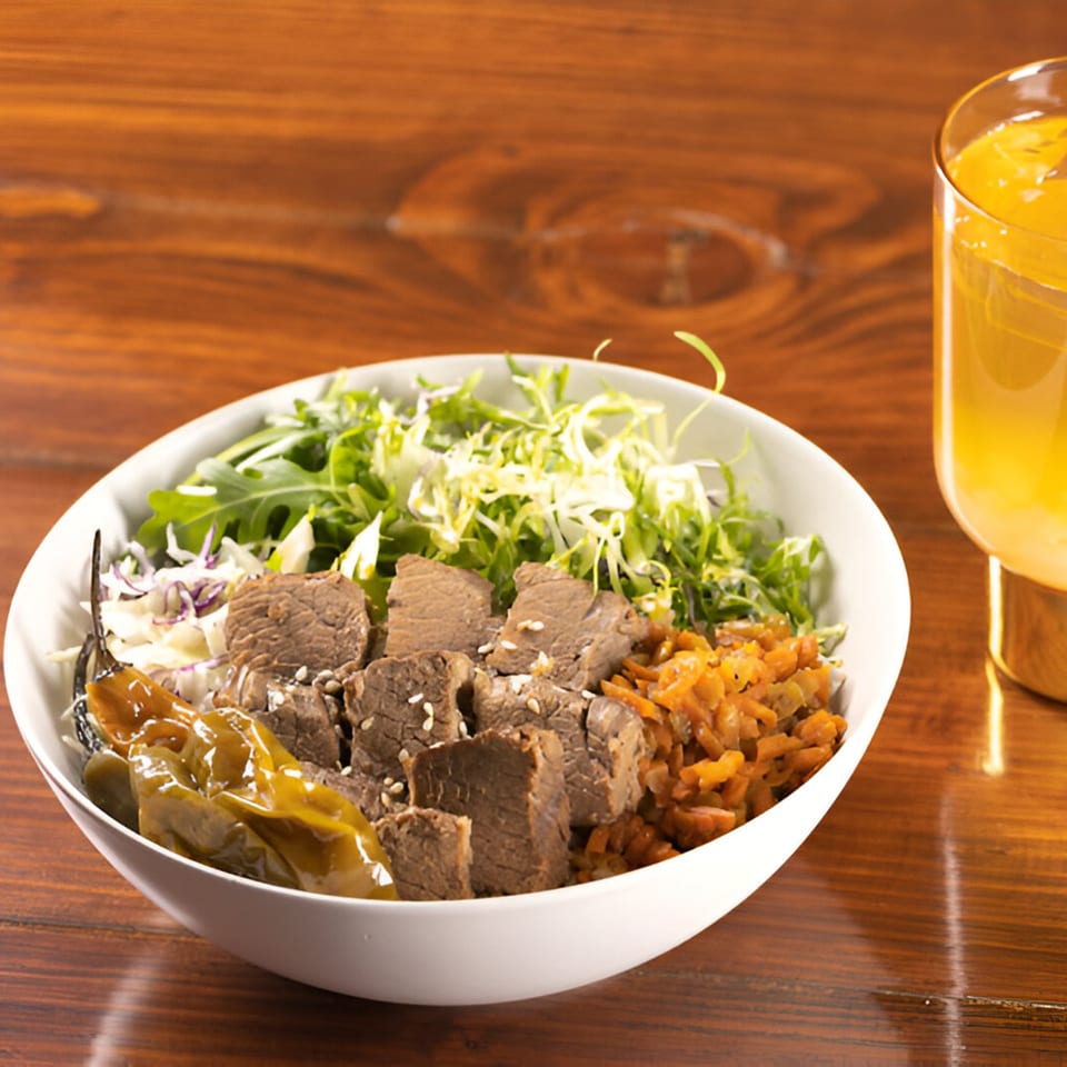 A Korean BBQ bowl with beef, greens, kimchi, and peppers, next to a glass of orange juice.