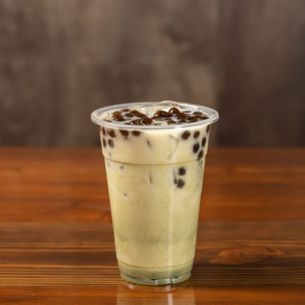 A plastic cup of light green boba tea with dark brown tapioca pearls and ice on a wooden table.