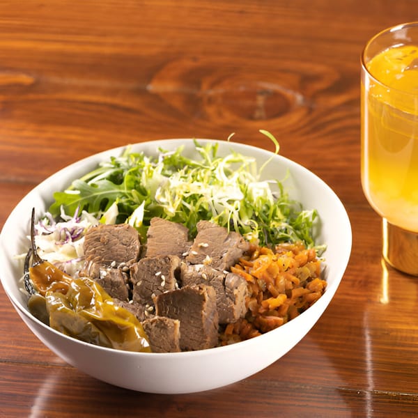 A white bowl of Korean BBQ beef, rice, greens, and peppers, with a drink beside it.