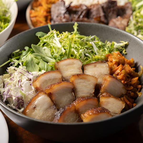 Close-up of a Korean BBQ dish with crispy pork belly, shredded cabbage, and greens in a dark bowl.