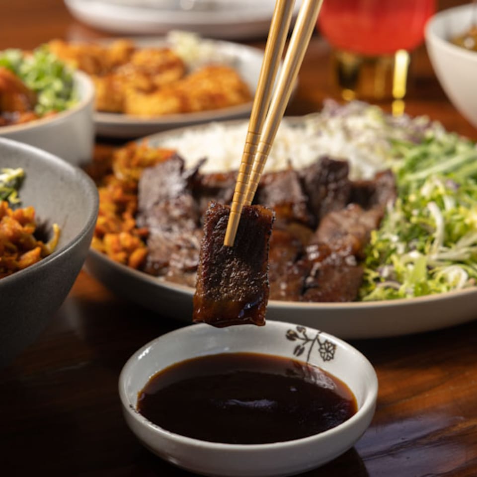Chopsticks dipping a piece of Korean BBQ beef into a dark sauce, with a meal in the background.